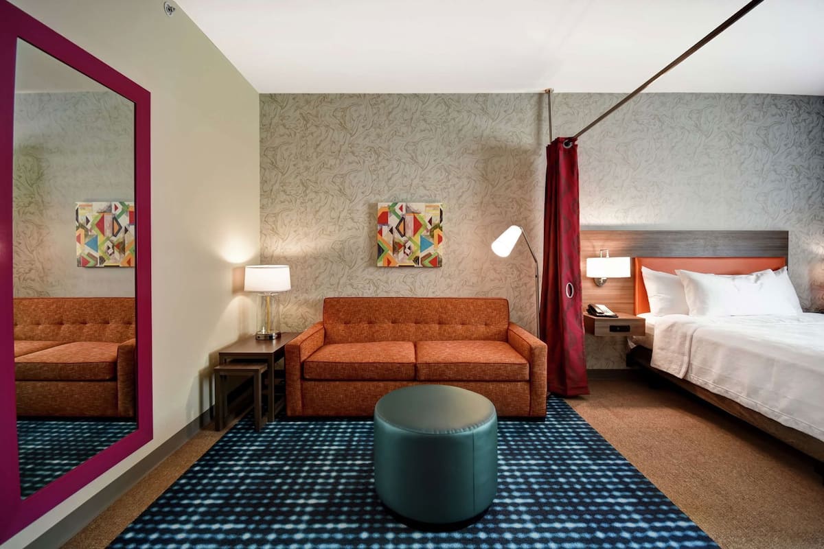 A hotel room with a sofa, a bed, a nightstand, a lamp, a mirror, and two wall art pieces.