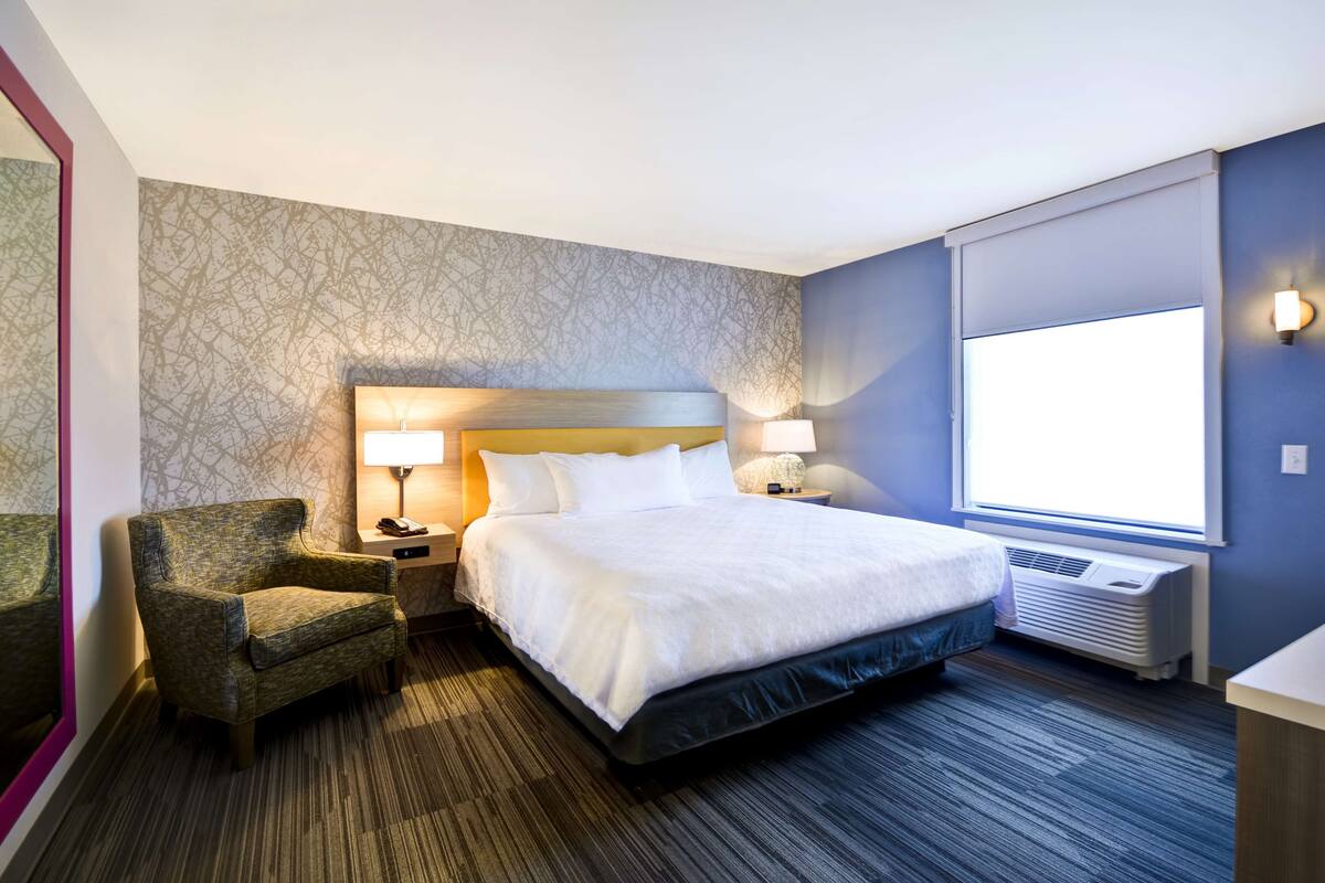 A hotel room with a large bed, a chair, a nightstand with a lamp, a wall-mounted air conditioner, and a window with daylight.