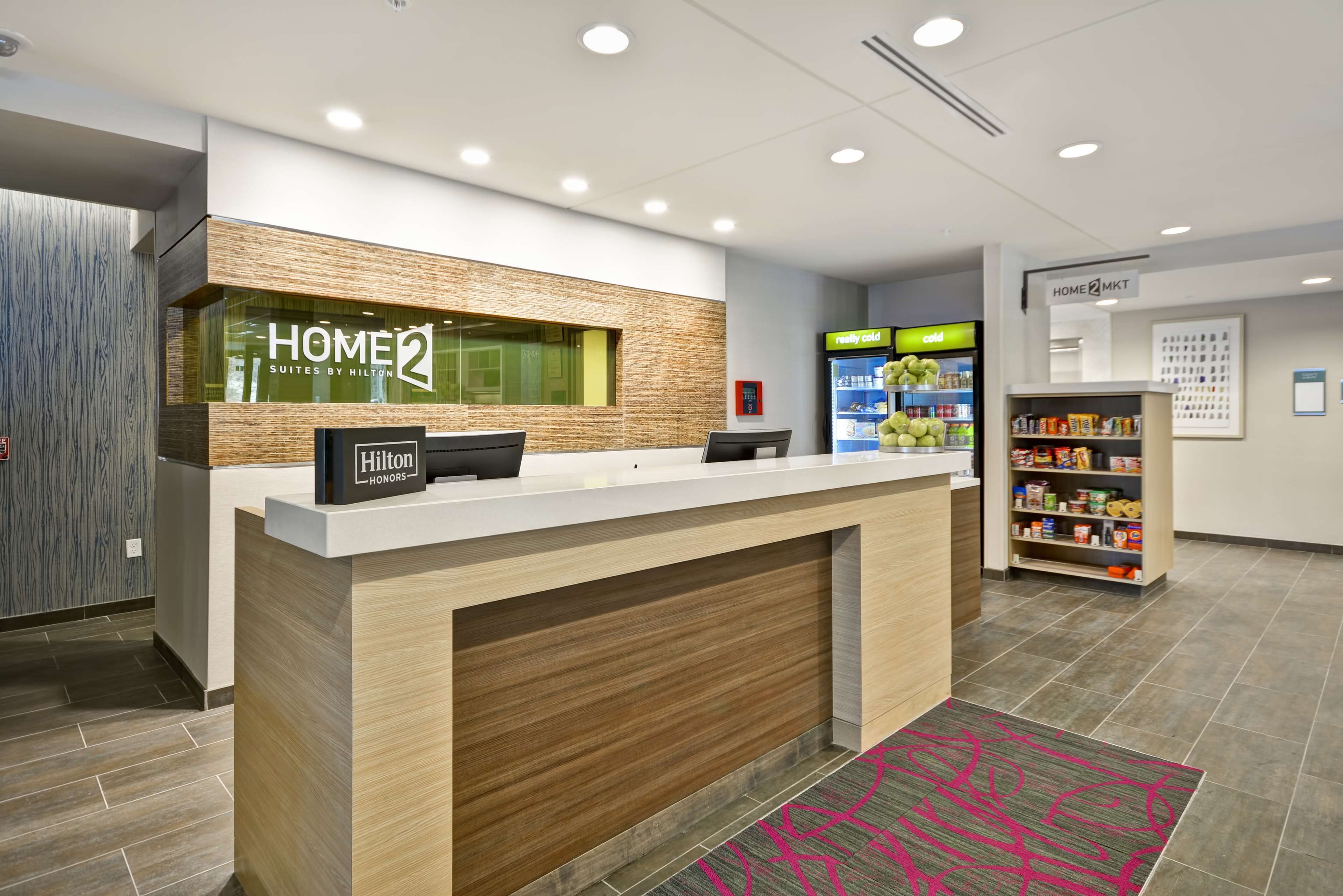 Zona de recepción de un hotel con un mostrador de recepción, un letrero de 'Home2 Suites by Hilton', un letrero de 'Hilton Honors', un refrigerador 'realmente frío' y un letrero de 'Home2 MKT'.
