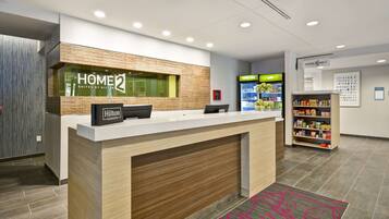 Zona de recepción de un hotel con un mostrador de recepción, un letrero de 'Home2 Suites by Hilton', un letrero de 'Hilton Honors', un refrigerador 'realmente frío' y un letrero de 'Home2 MKT'.