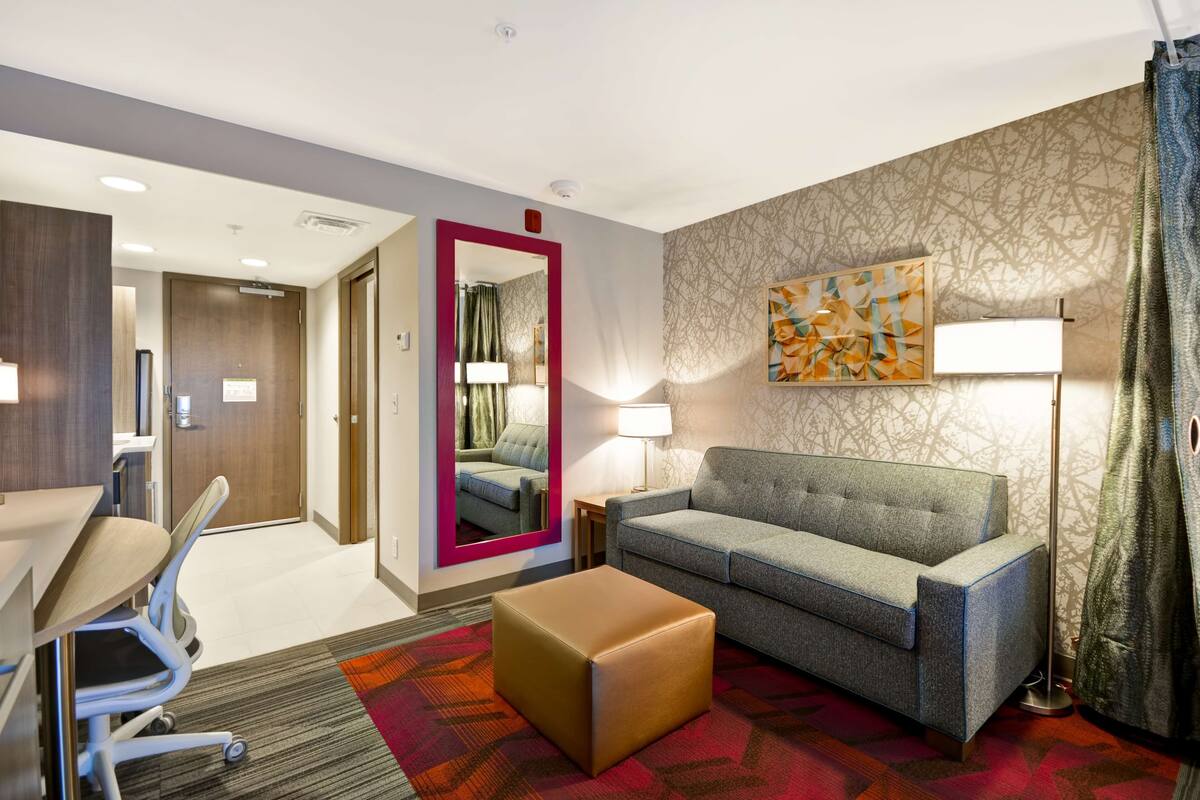 A hotel room with a sofa, ottoman, desk, chair, and a mirror.