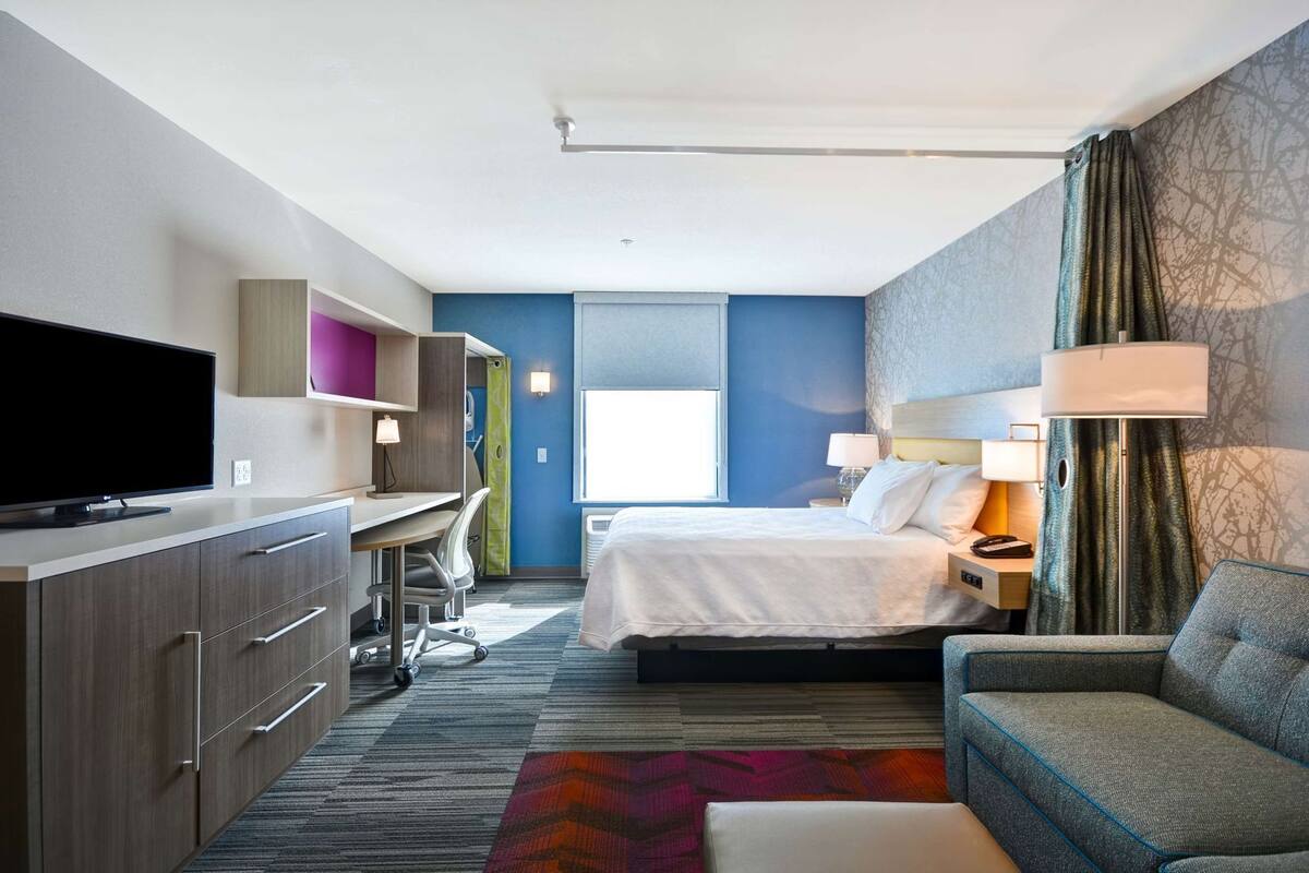 A hotel room with a bed, a desk with a TV, a sofa, and a window.