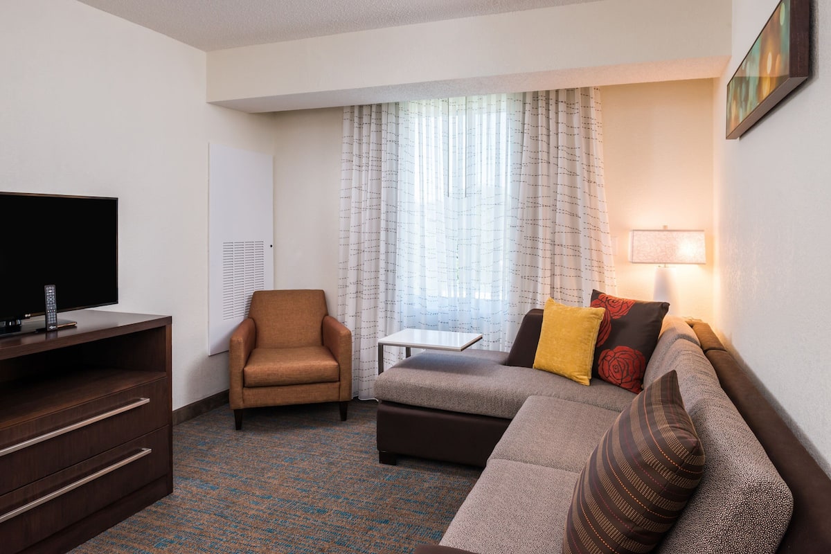 A hotel room with a flat-screen TV, a brown armchair, a gray sofa with patterned cushions, a small table, and a lamp.