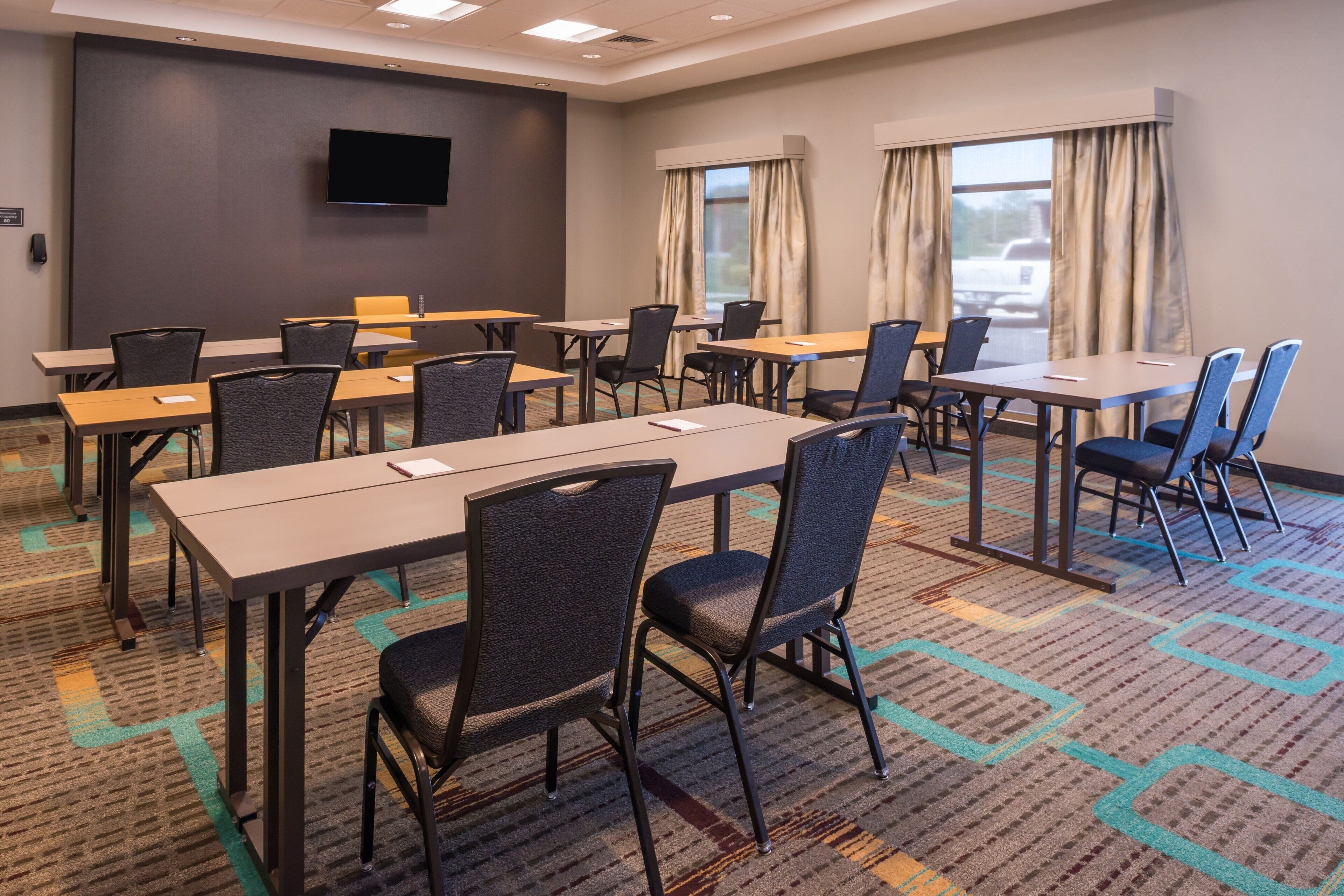 A conference room with multiple tables and chairs, a flat-screen TV mounted on the wall, and large windows with curtains.