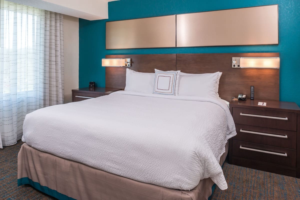 A hotel room with a large bed, a wooden headboard, two bedside tables, and a view of the outside through a window with curtains.