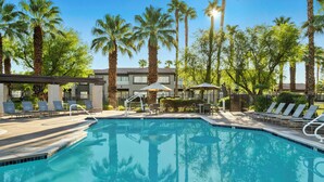 A swimming pool with lounge chairs and umbrellas, surrounded by palm trees and a building with a tiled roof. - Worldmark Cathedral City (Cathedral City)