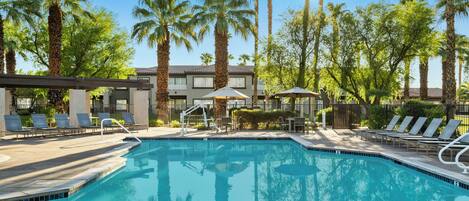 A swimming pool with lounge chairs and umbrellas, surrounded by palm trees and a building with a tiled roof.