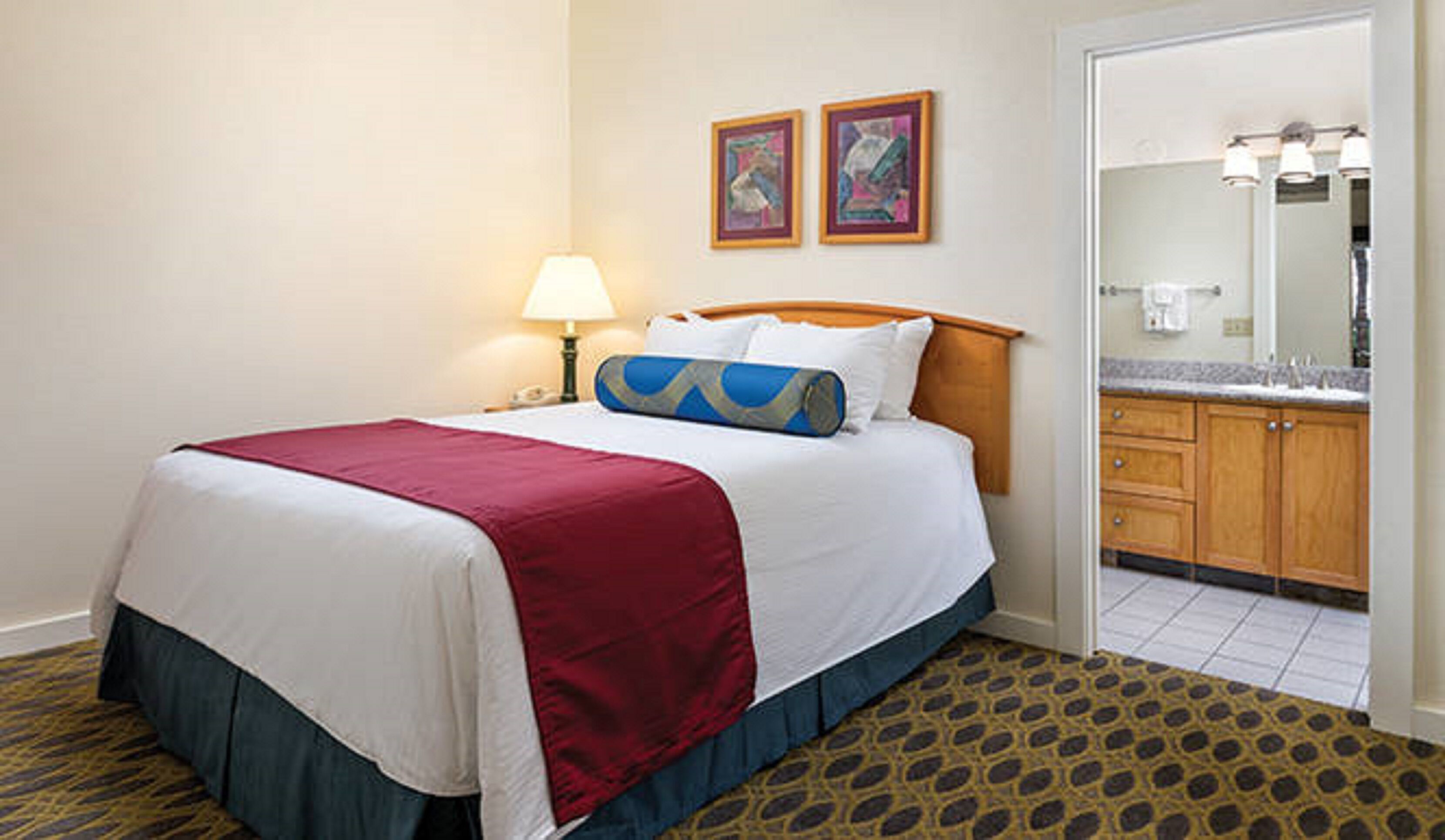 A hotel room with a bed, a nightstand, a lamp, a bathroom with a sink and mirror, and two framed pictures on the wall.