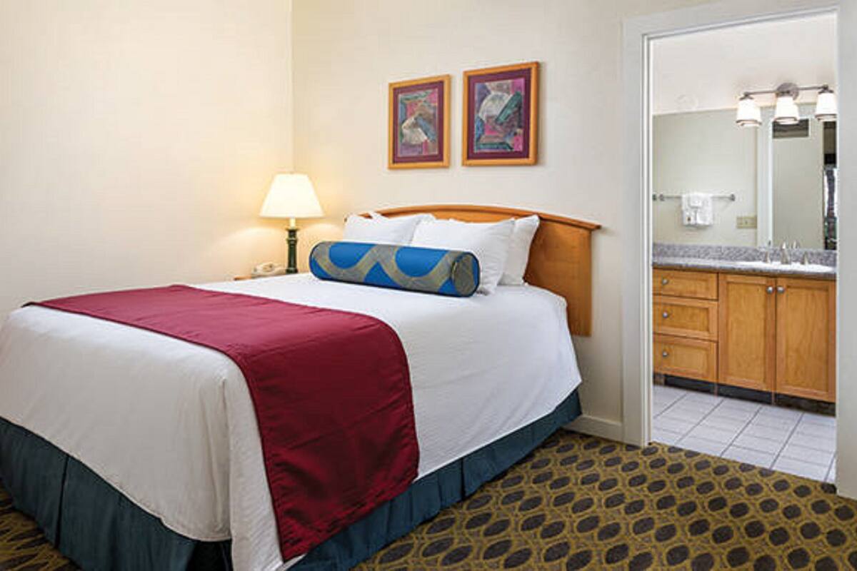A hotel room with a bed, a nightstand, a lamp, a bathroom with a sink and mirror, and two framed pictures on the wall.