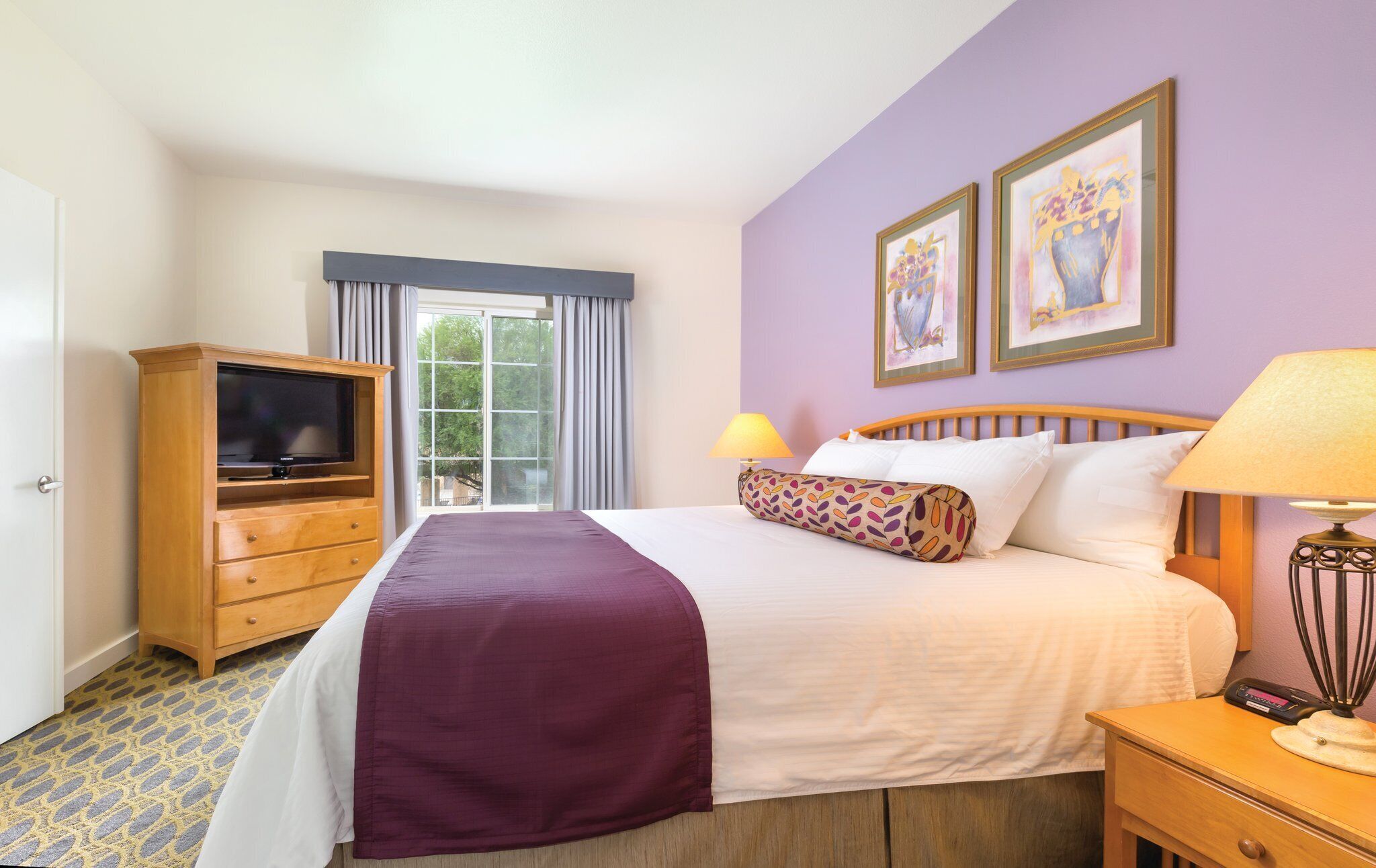 A hotel room with a bed, a television, a desk with a lamp, and two framed artworks on the wall.