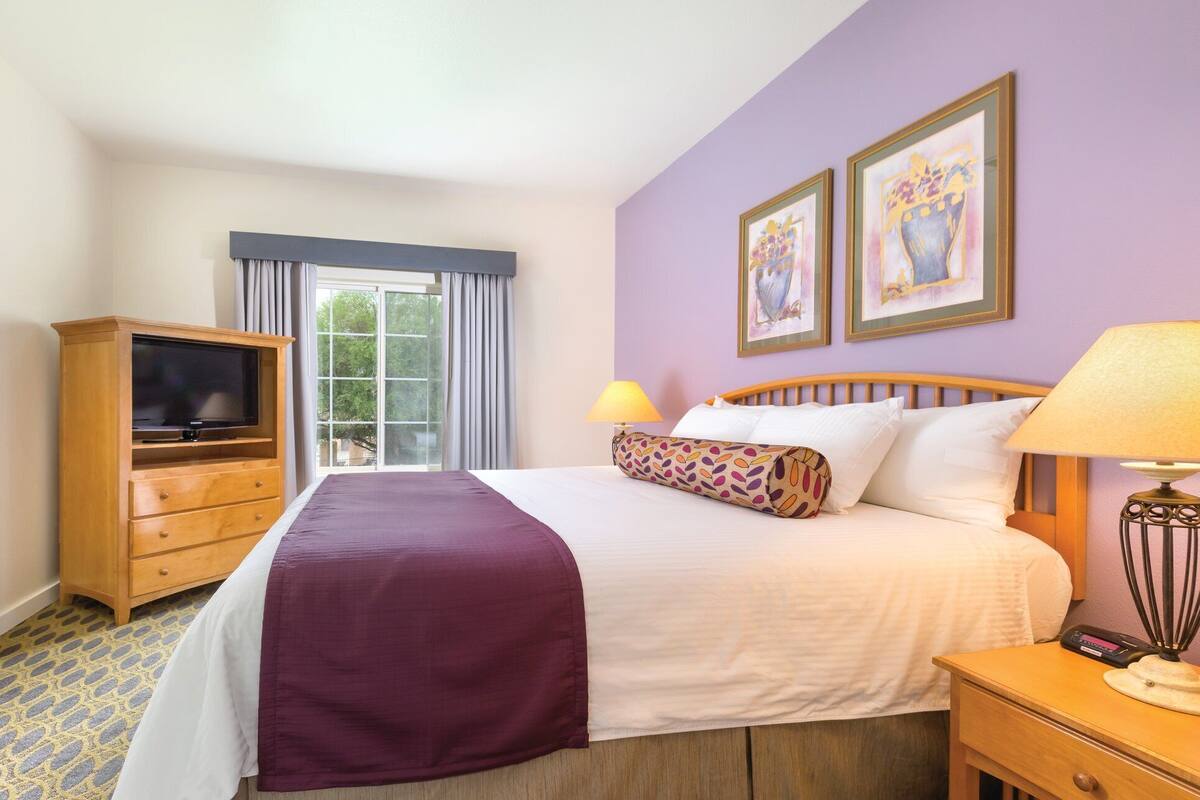 A hotel room with a bed, a television, a desk with a lamp, and two framed artworks on the wall.