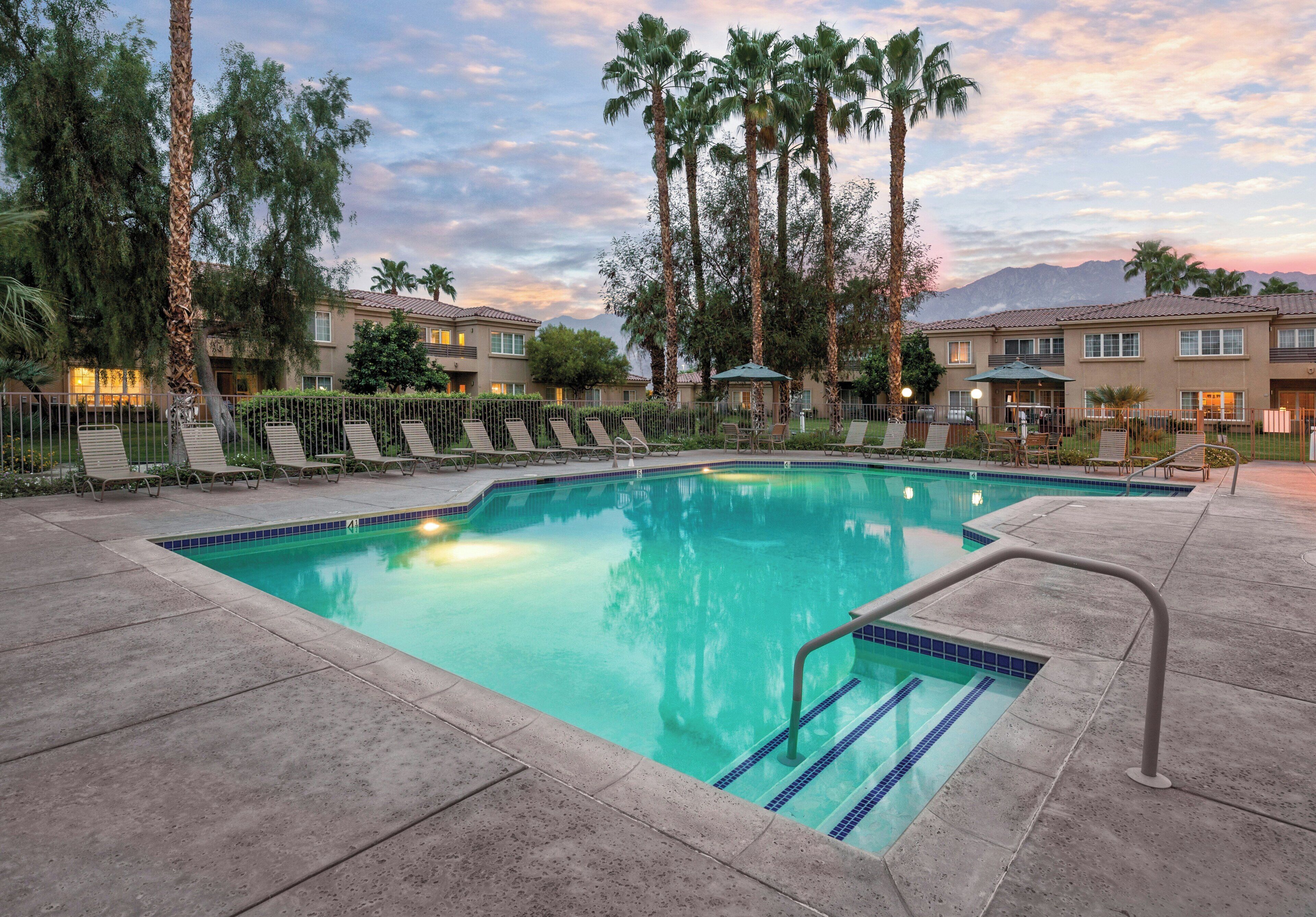 A swimming pool with lounge chairs and palm trees, surrounded by buildings with a view of mountains.