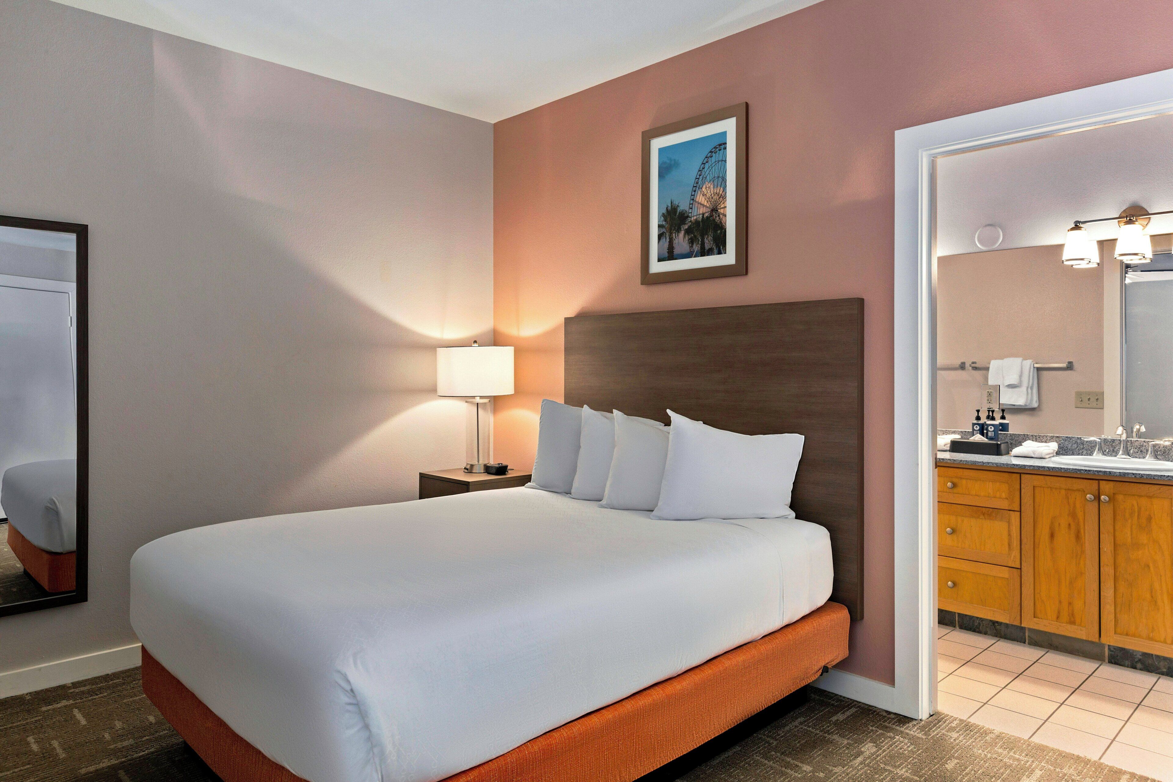 Une chambre d’hôtel avec un grand lit, une table de chevet avec une lampe, une salle de bain avec un lavabo et un miroir, et un tableau encadré au mur.
