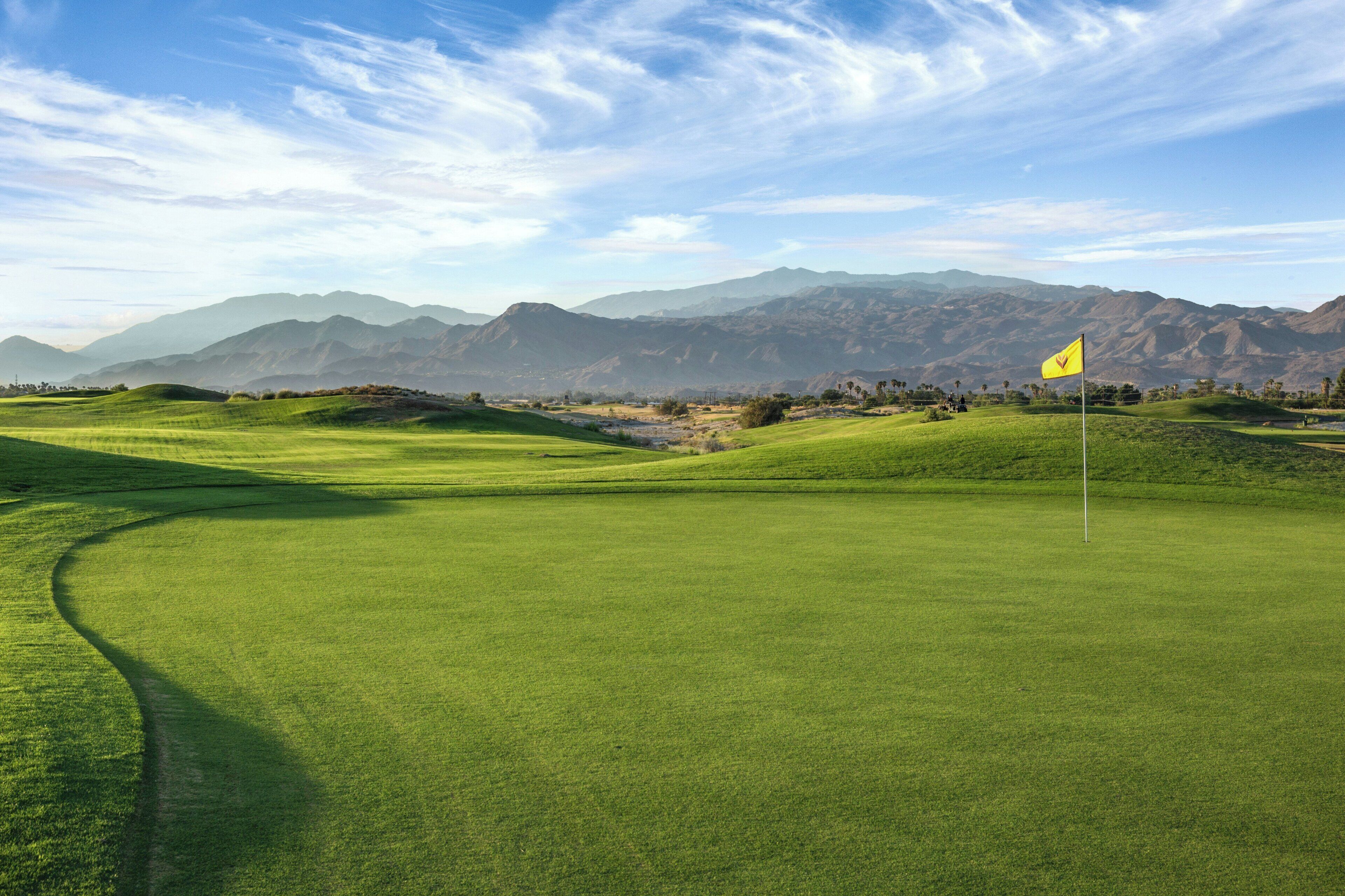 A golf course with a flag, rolling hills, and mountains in the background.