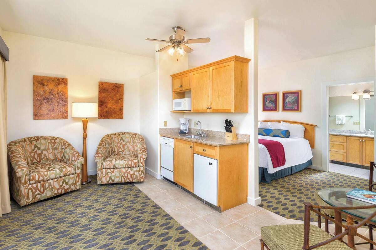 A compact hotel room with a kitchenette, two patterned armchairs, a bed with a red blanket, and a dining area with a glass table.