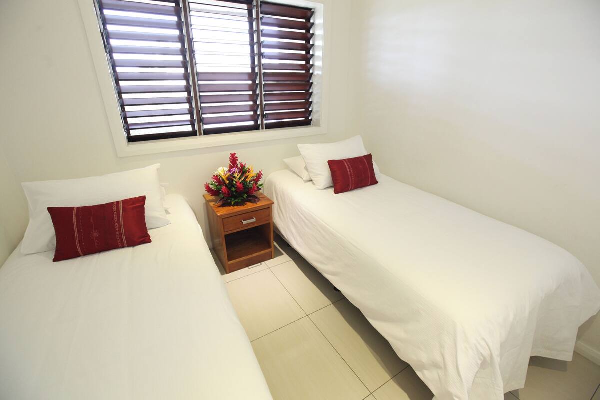 Habitación de hotel con dos camas individuales, una mesita de noche de madera con un florero y ropa de cama blanca, además de una ventana con persianas.