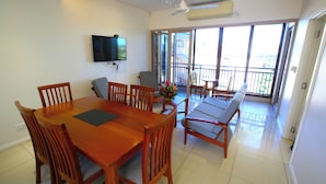 A dining area with a wooden table and chairs, a flat-screen TV mounted on the wall, and a balcony with a view of the outdoors. - Premiere Apartments (Nadi)