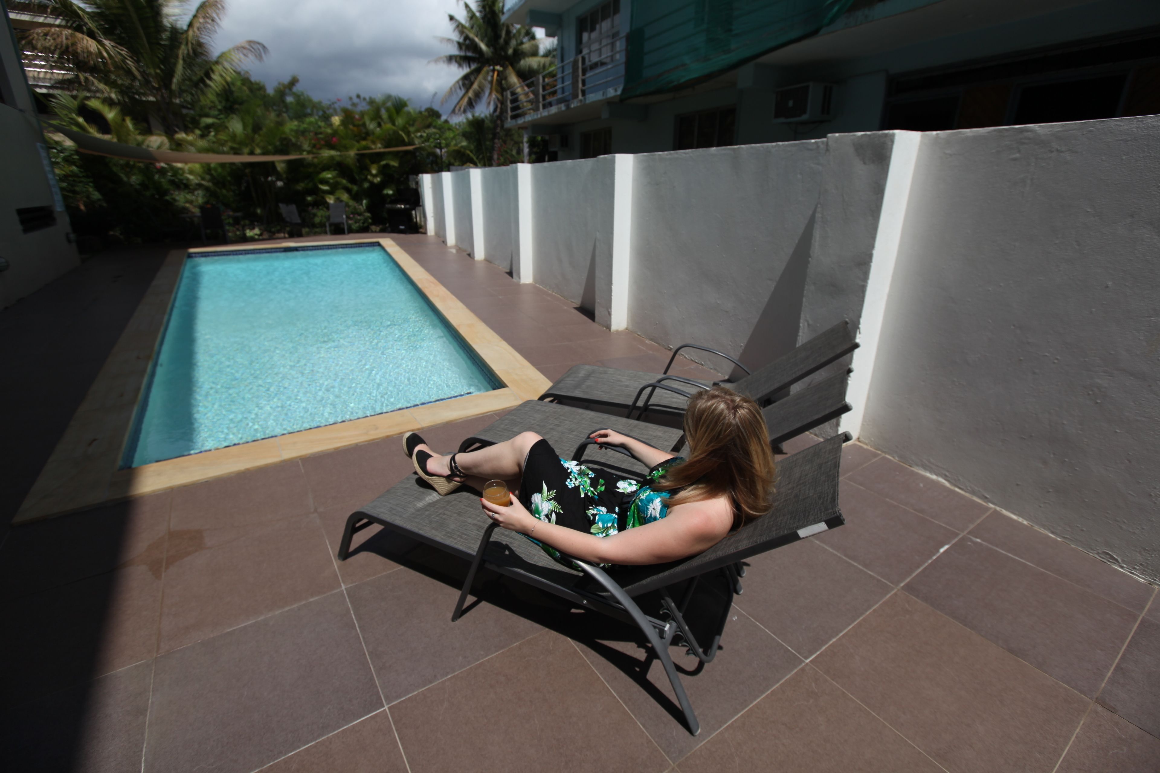 Una mujer descansando en una silla de lounge junto a una piscina, con una pared blanca y palmeras al fondo.