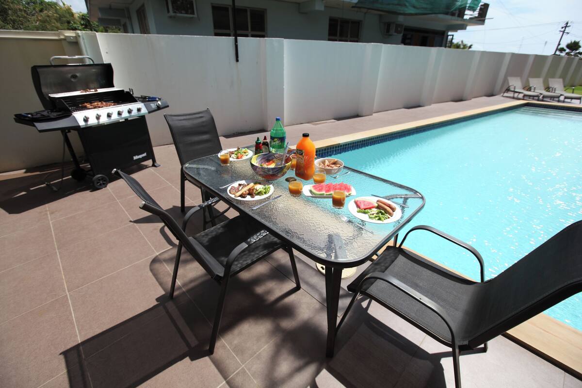 Un espacio exterior para comer con una mesa lista para dos personas, una parrilla y vista a la piscina.