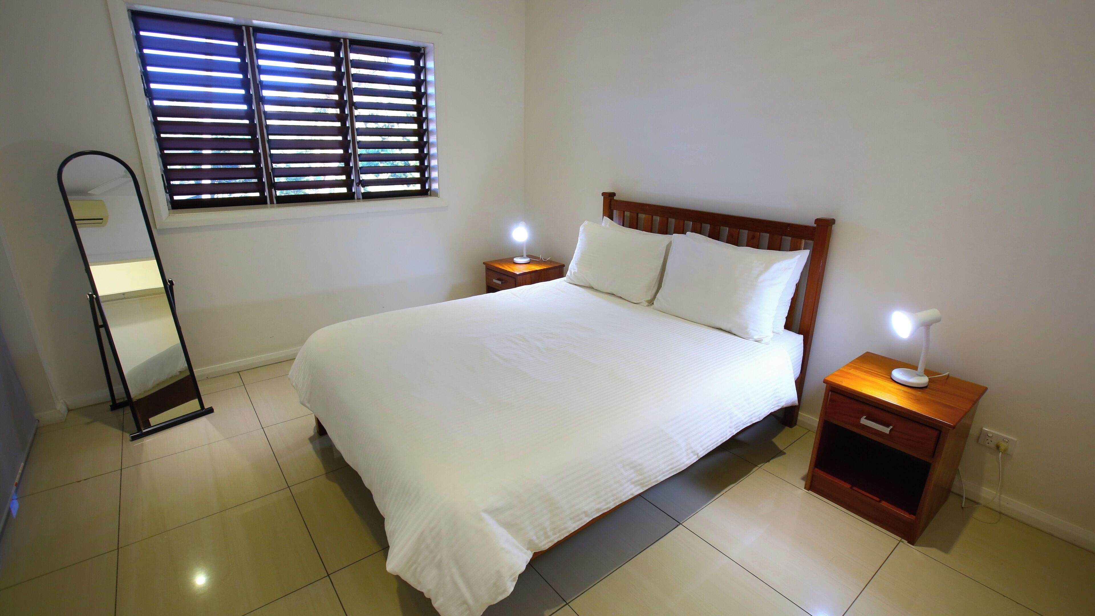 Una cama bien hecha con sábanas blancas, dos lámparas de noche, una mesita de madera, un espejo y una ventana con persianas.