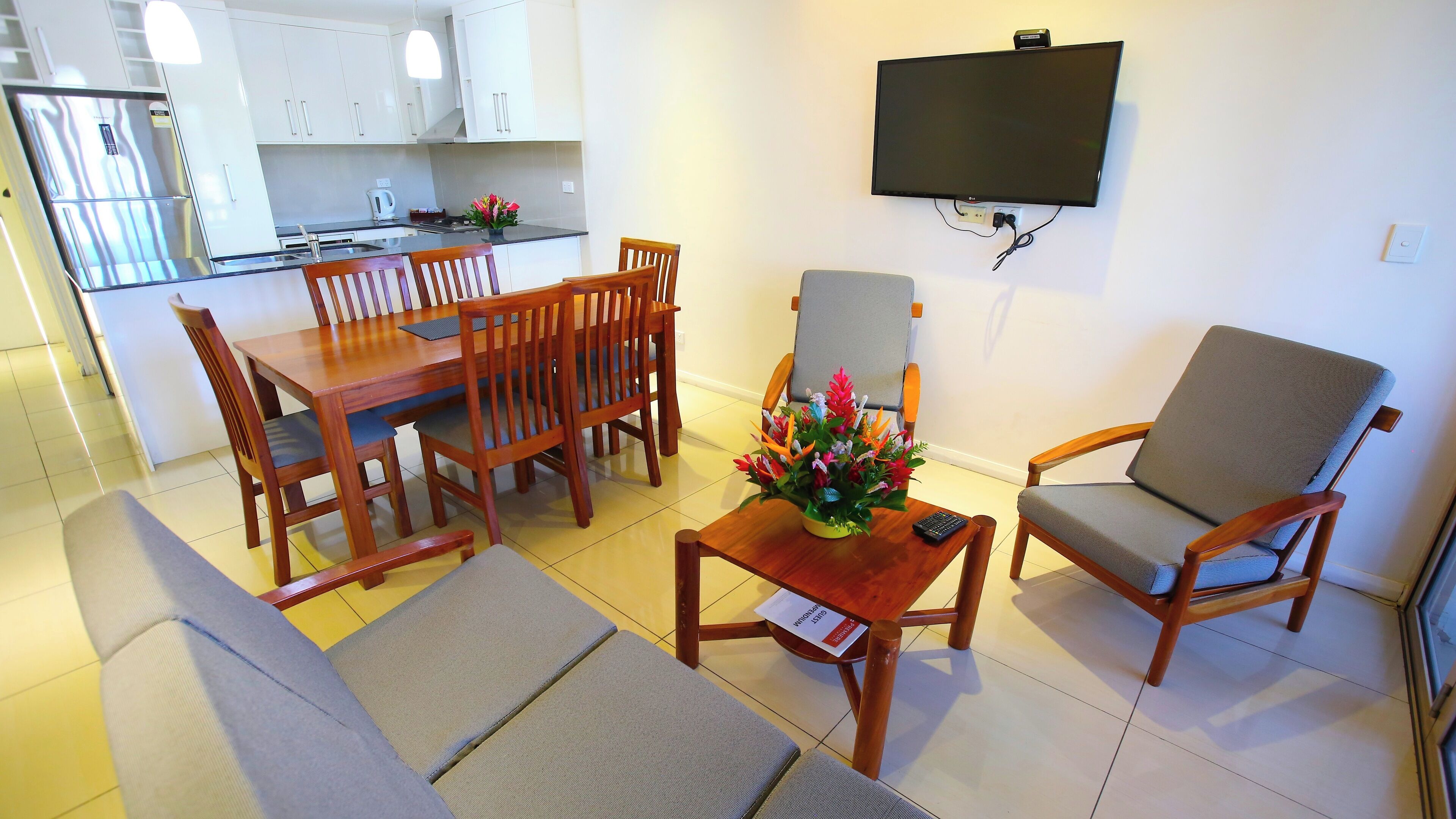 A modern kitchen with white cabinets, a dining table with wooden chairs, a grey sofa, and a grey armchair. A flat-screen TV is mounted on the wall.
