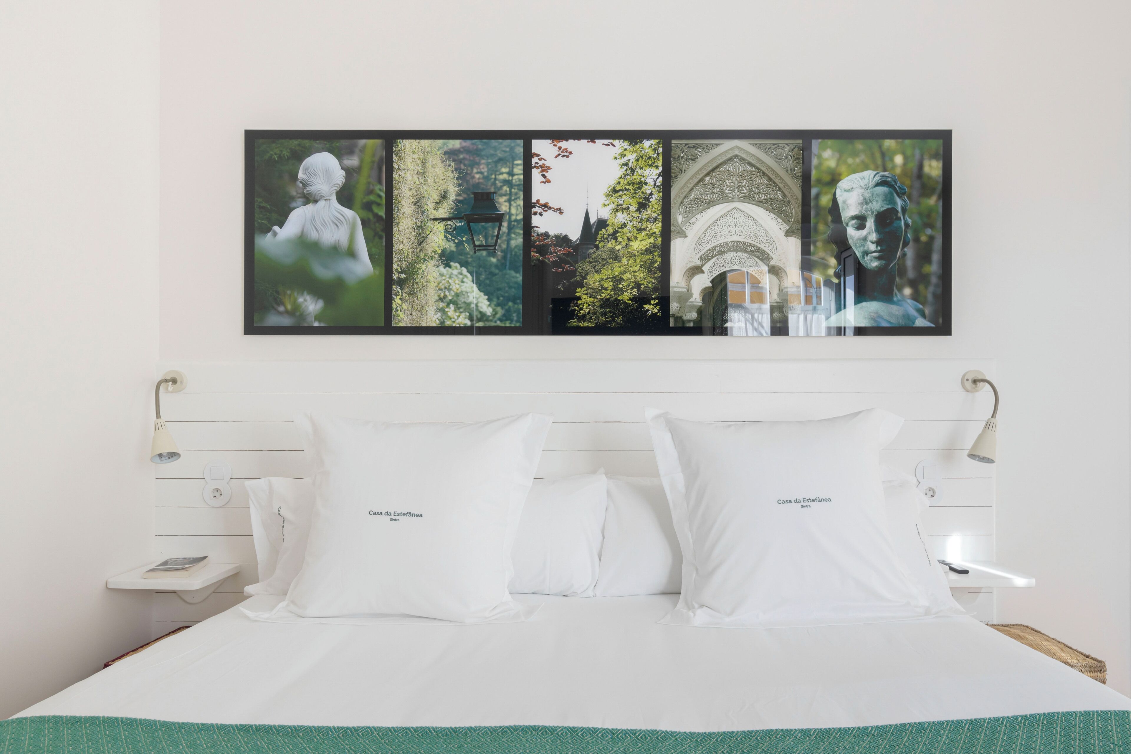 Una cama bien hecha con sábanas y almohadas blancas, un cuadro enmarcado sobre la cama y una mesita de noche sencilla con una lámpara.