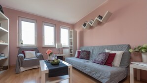 Ein modernes Wohnzimmer mit einem grauen Sofa, einem weißen Couchtisch und einem grauen Sessel. Zwei Fenster mit weißen Rahmen, ein weißes Bücherregal und ein weißer Beistelltisch mit einem Buch sind ebenfalls vorhanden.