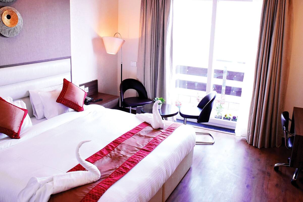 A hotel room with a large bed, a desk, a chair, a lamp, and a view of the outside.