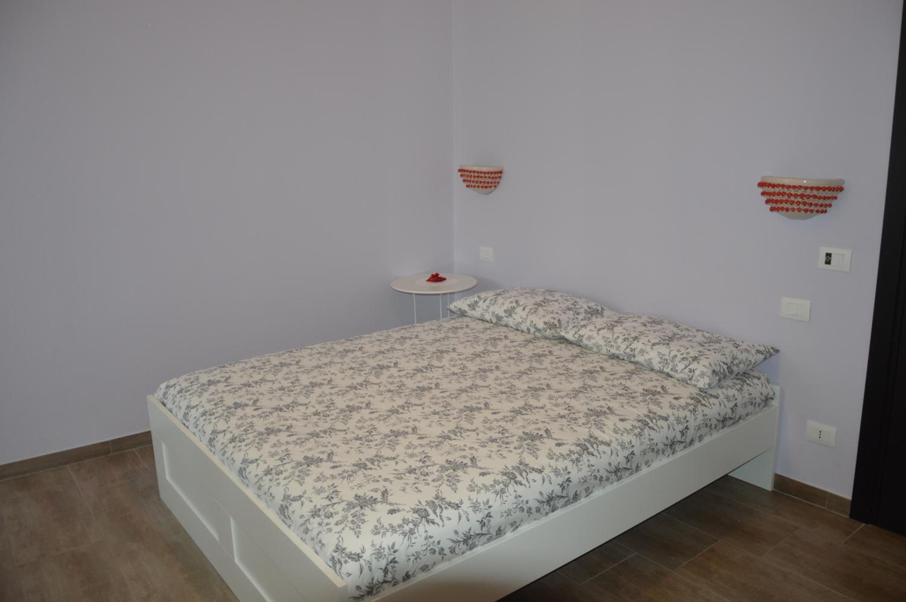 Una cama individual con una colcha estampada en una habitación con paredes de colores claros y una pequeña mesa redonda con un objeto rojo encima.