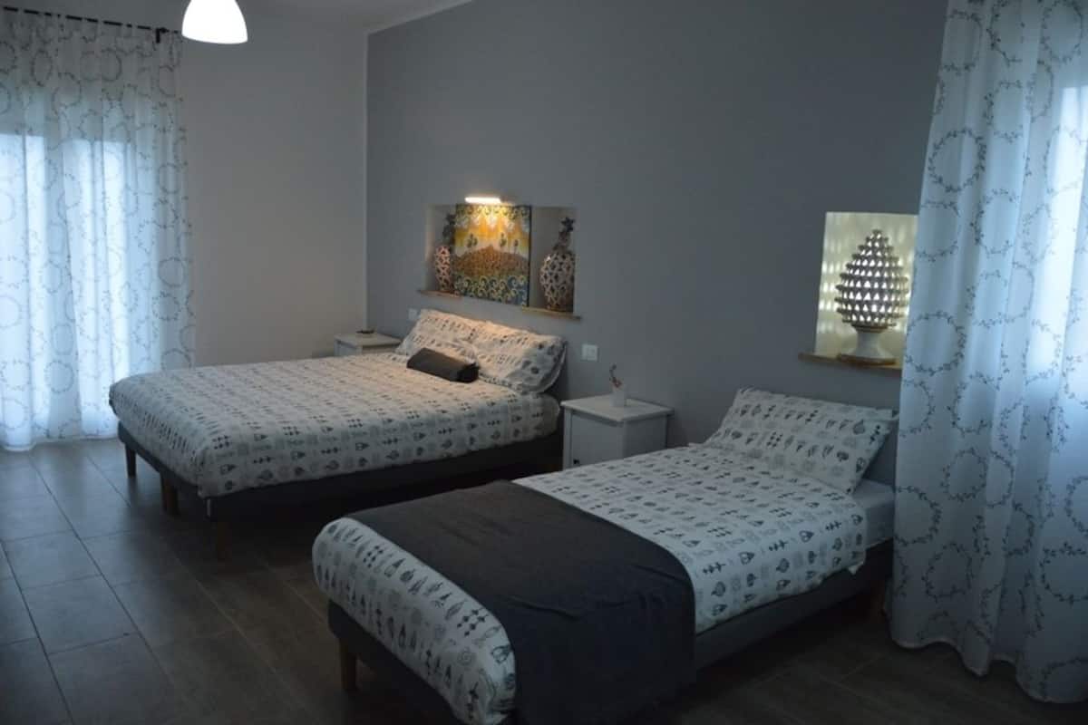 A bedroom with two beds, a nightstand, a wall-mounted shelf with decorative items, and a window with a patterned curtain.