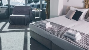 Hotelkamer met een groot bed, een stoel, een klein tafeltje met planten en uitzicht op de stad door grote ramen.