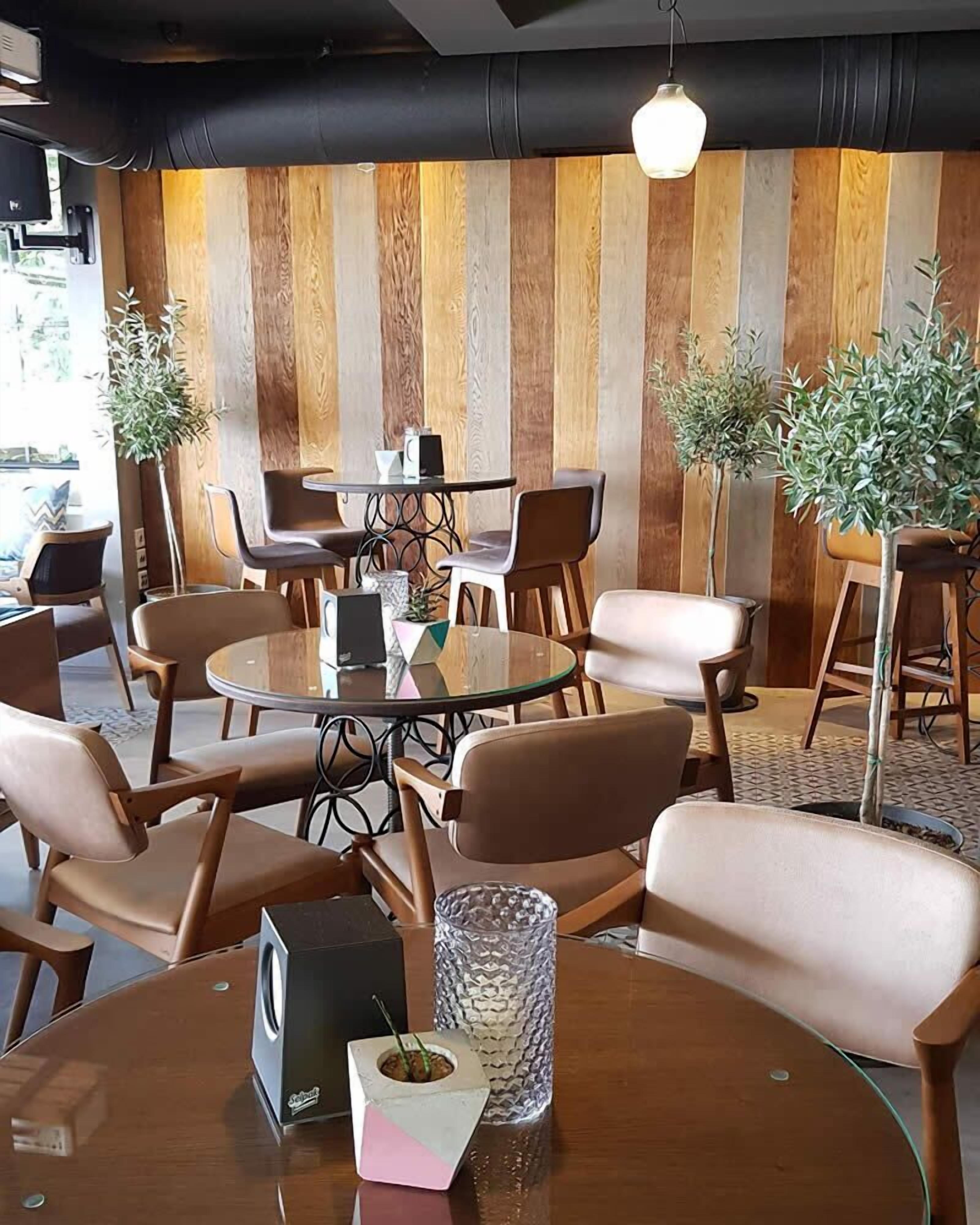 A modern dining area with round tables and cushioned chairs, wooden panel walls, and potted plants.