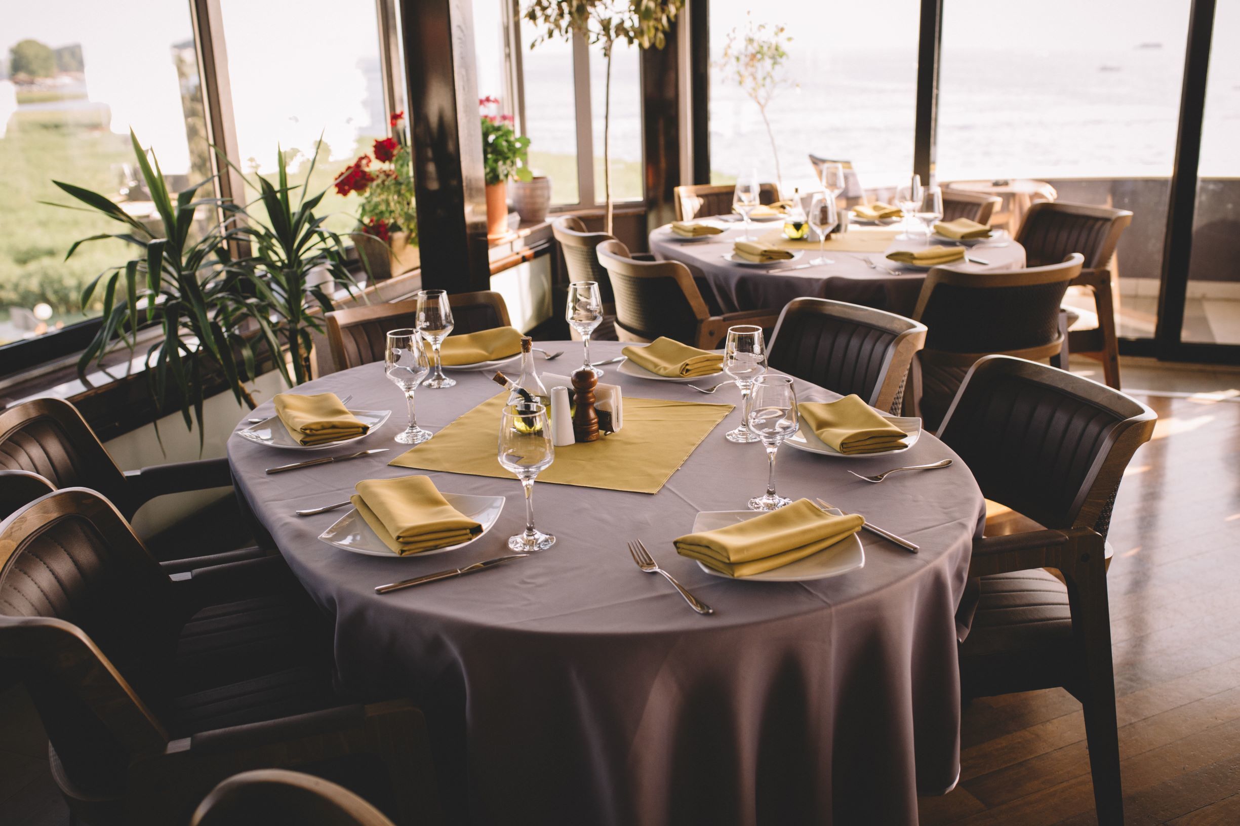 Un espace repas avec des tables rondes disposées pour accueillir quatre personnes, chacune étant recouverte d’une nappe et agrémentée d’assiettes, de couverts et de verres. Des chaises sont placées autour des tables, et la pièce est dotée de grandes fenêtres offrant une vue sur l’extérieur.
