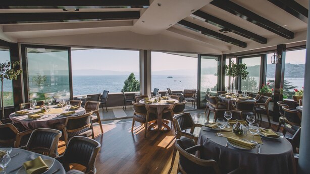 Een restaurant met meerdere tafels en stoelen, grote ramen met uitzicht op zee, en een modern interieur.