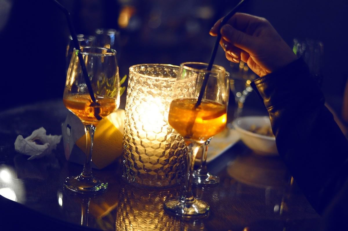 Un bar con vasos de bebidas de color ámbar, una vela con textura y una mano removiendo una bebida.