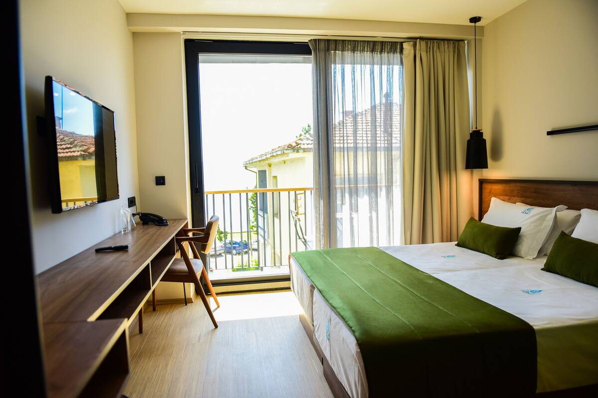 A hotel room with a bed, a desk, a chair, a TV, and a balcony with a view of buildings.