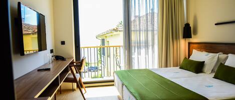 A hotel room with a bed, a desk, a chair, a TV, and a balcony with a view of buildings.