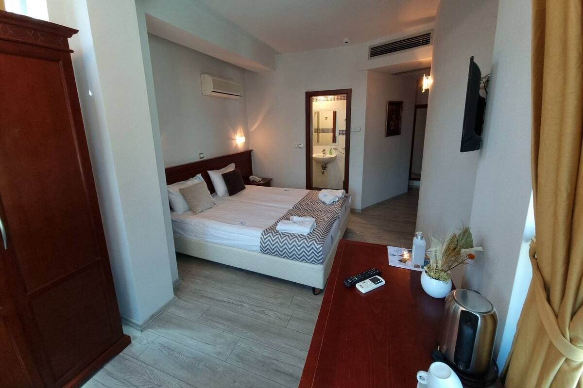 A hotel room with a bed, bedside table, and a small desk with a kettle and a plant.