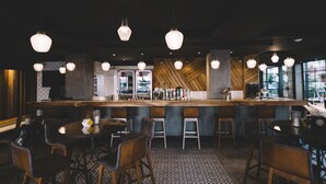 Een bar met hangende lampen, een houten toog, barkrukken en een vloer met een patroon.