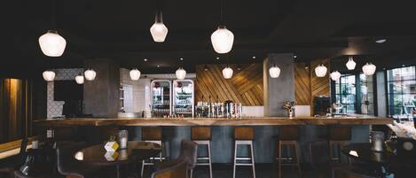 A bar area with hanging light fixtures, wooden bar counter, stools, and a patterned floor.