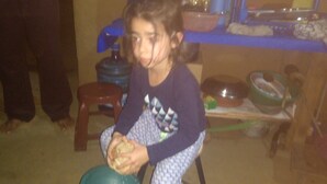 Uma criança sentada num banquinho em um quarto com um bebedouro, uma tigela e uma prateleira com diversos objetos.