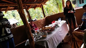 Uma área de refeições ao ar livre coberta, com mesa posta para uma refeição, rodeada por mobiliário de madeira e vegetação.