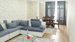 Ein Wohnzimmer mit Ecksofa, Glastisch, Esstisch mit Stühlen und gemusterten Teppichen.