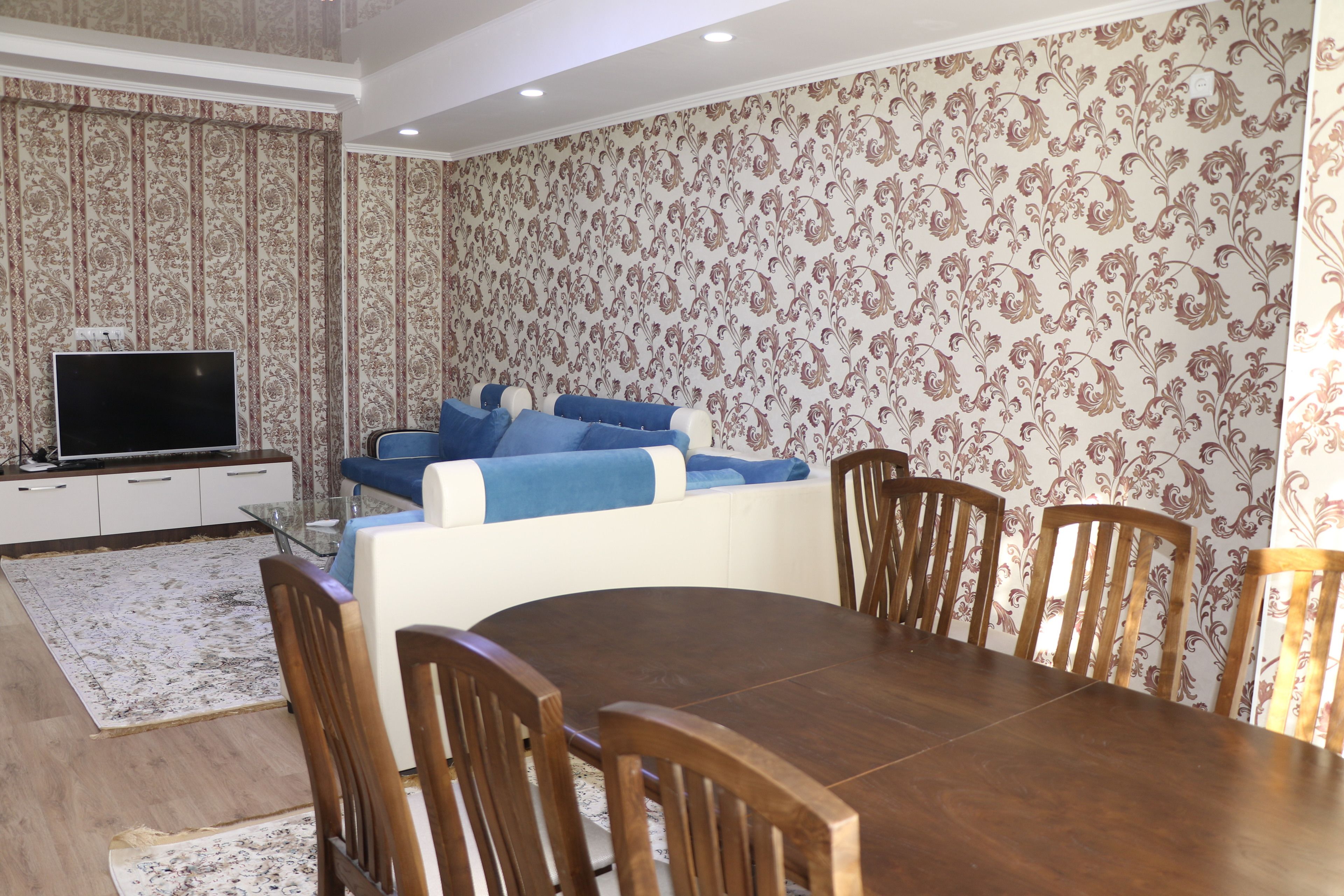 Un comedor con una mesa y sillas de madera, un sofá con cojines azules y un televisor sobre un mueble blanco.