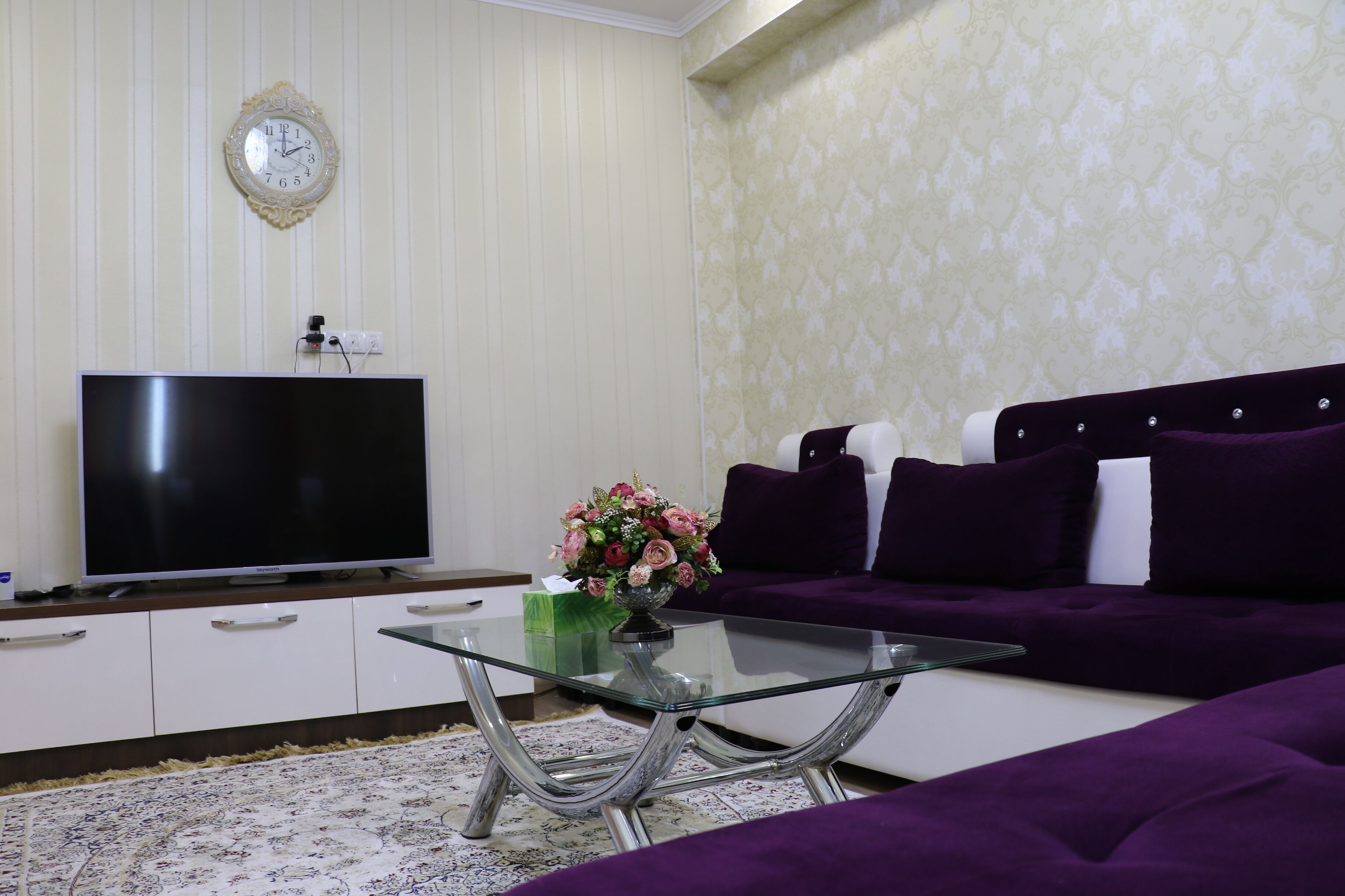 Un salón con televisor de pantalla plana, una mesa de centro de vidrio, un sofá púrpura y un tapete estampado.