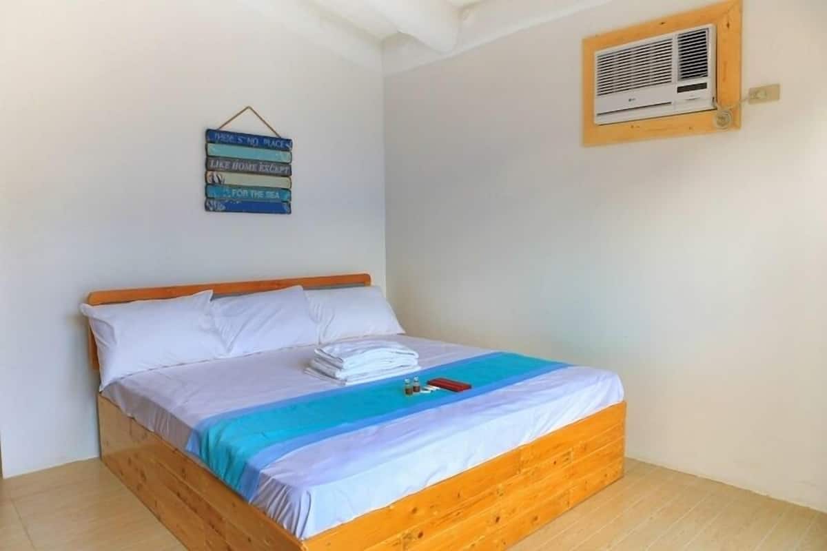 Una cama bien hecha con sábanas blancas y una manta azul y blanca a rayas, una mesita de noche con un objeto rojo y un aire acondicionado instalado en la pared.
