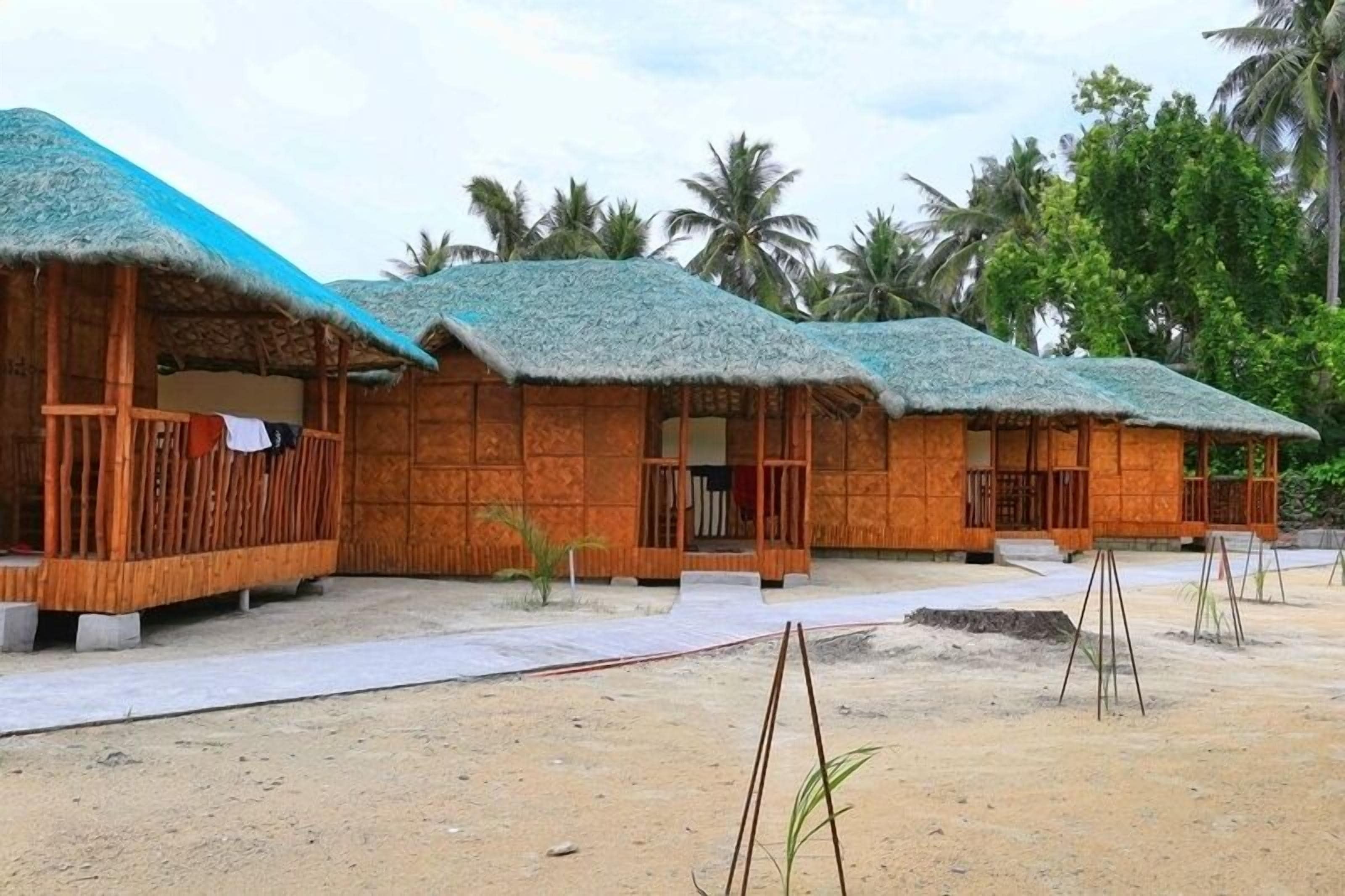 A row of thatched-roof huts with wooden walls, a sandy ground, and palm trees in the background.