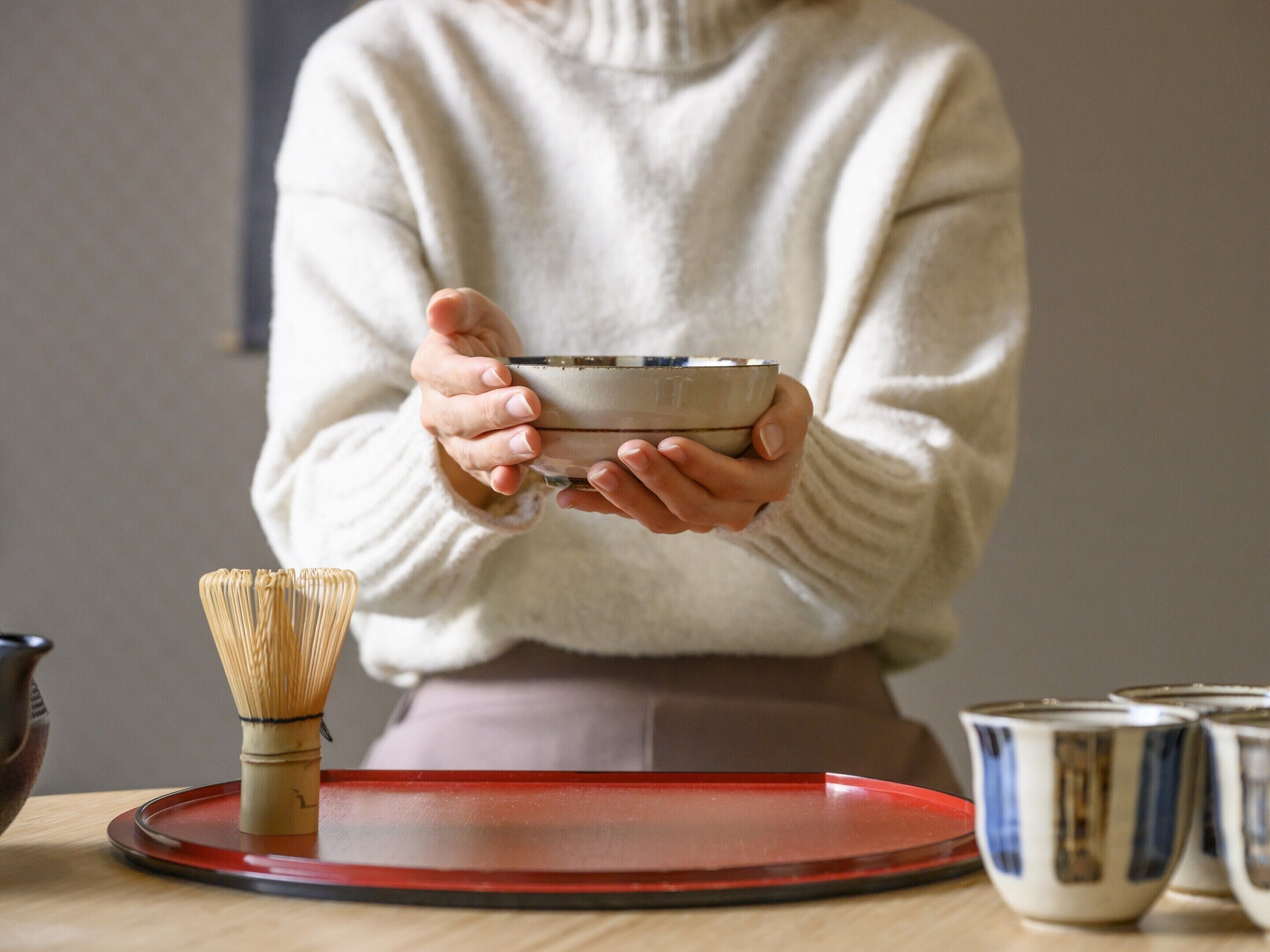 Une personne tenant un saladier, un fouet et un plateau rouge avec des tasses, le tout posé sur une table.