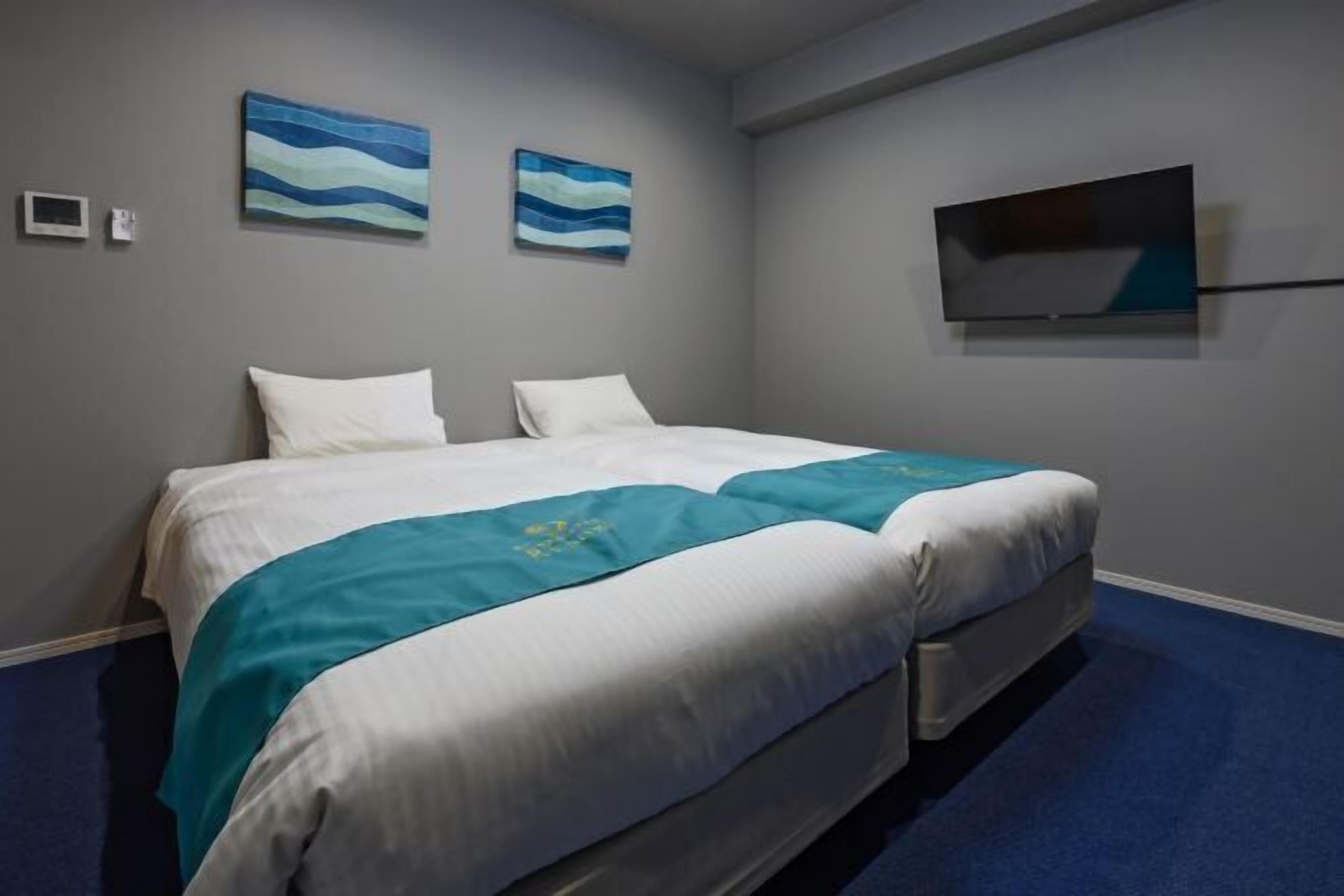 A hotel room with a bed, two pillows, a blue carpet, a television, and two wall-mounted paintings.