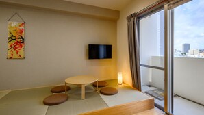 A room with tatami flooring, a low table, cushions, a TV, and a window with a city view. - TOKI NO YADO NAHA by SUNREST RESORT (Naha)